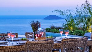 A dining table set with wine glasses, plates, and a bottle, overlooking a body of water with a distant island. - Eco Villa Mirada (Kalkan)