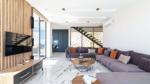 A modern living room with a large TV, a wooden entertainment unit, a grey sectional sofa, a coffee table, and a staircase leading to an outdoor area. - Eco Villa Mirada (Kalkan)