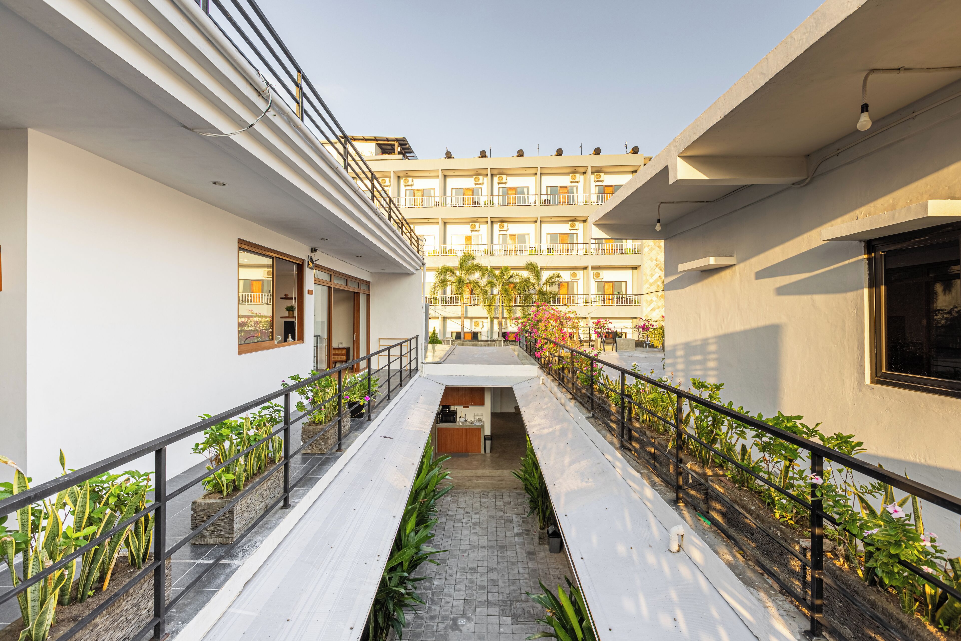 A corridor with a central walkway flanked by planters and railings, leading to a multi-story building with balconies.
