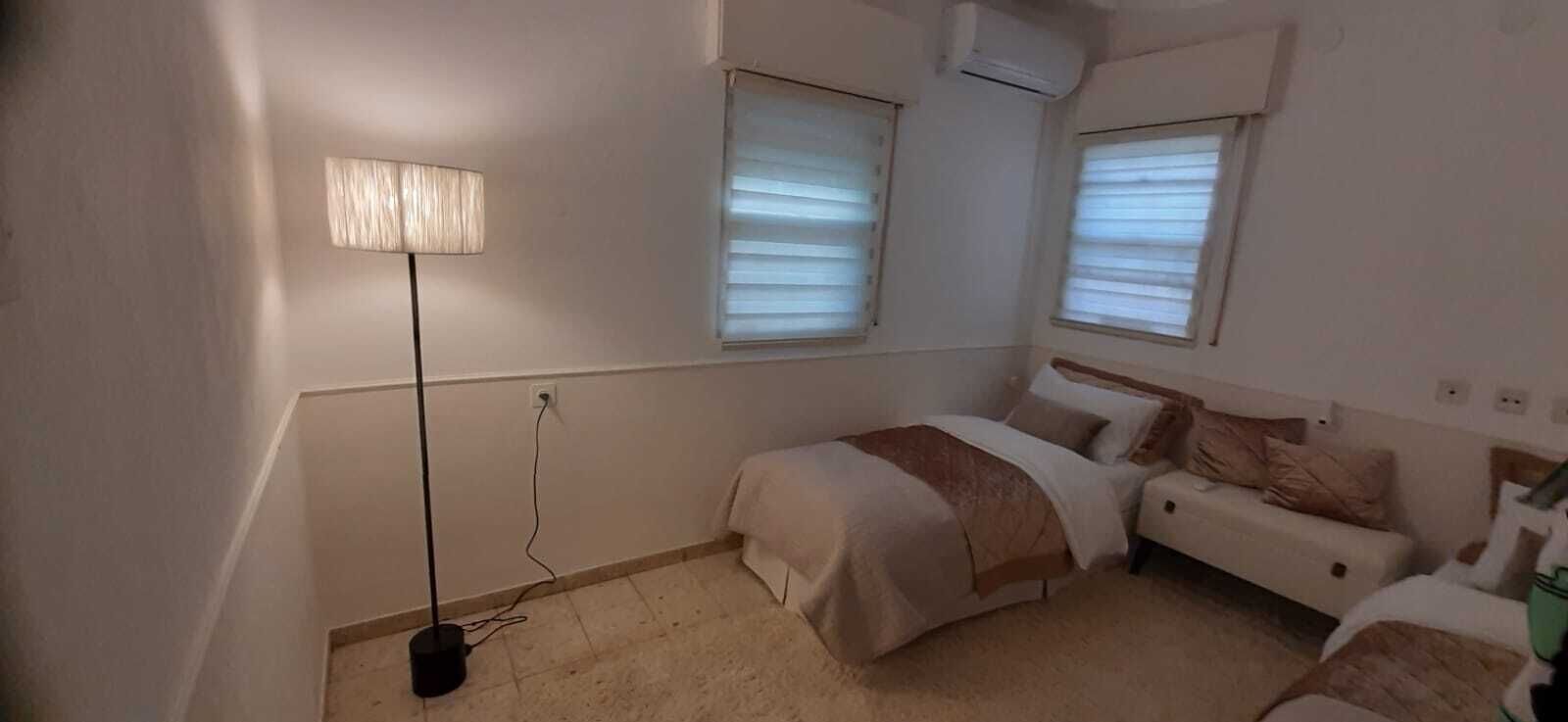 A small, neatly arranged bedroom with two beds, a bedside table, a lamp, and a window with blinds.
