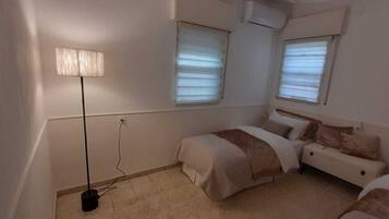 A small, neatly arranged bedroom with two beds, a bedside table, a lamp, and a window with blinds.