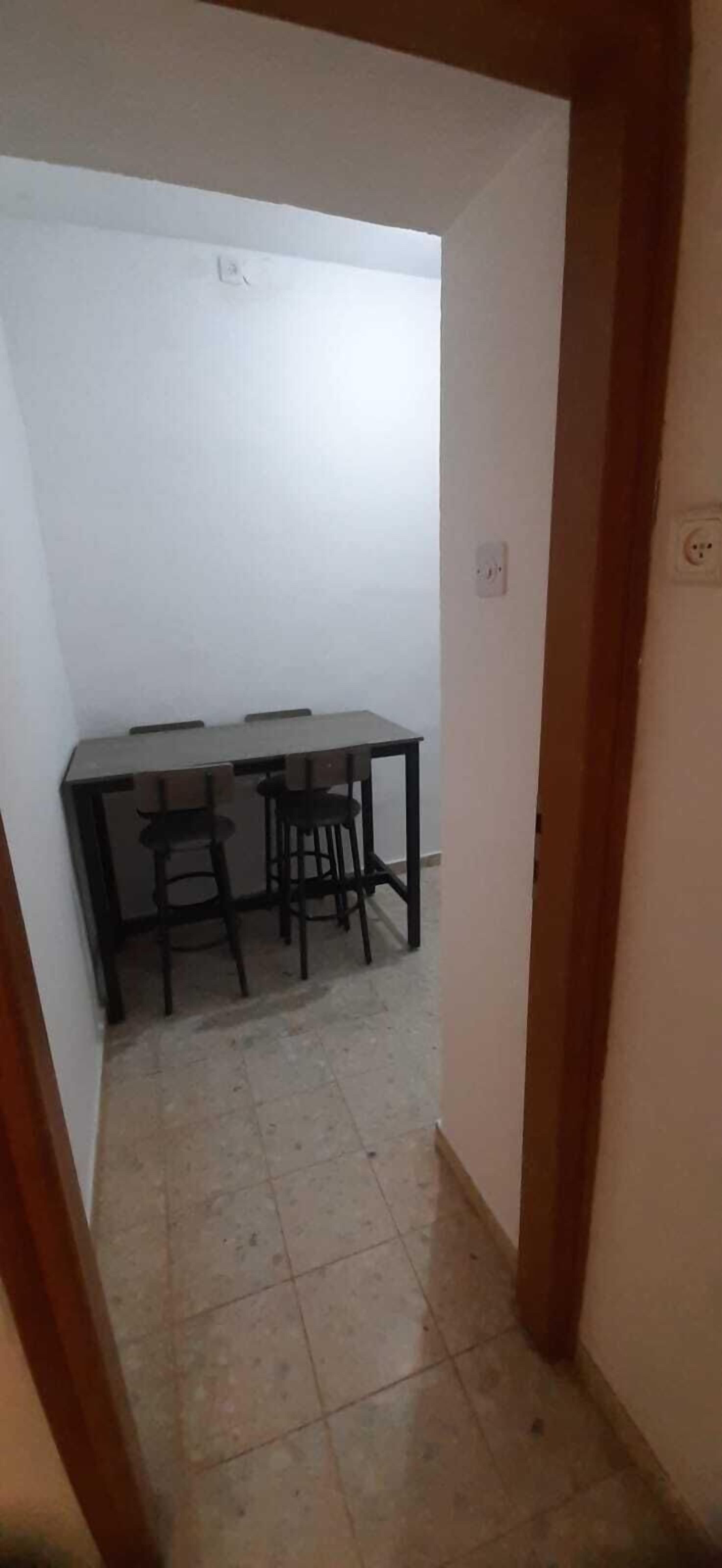 Passadís estret que condueix a una habitació amb una taula i caires, un terra de rajola i una paret blanca.