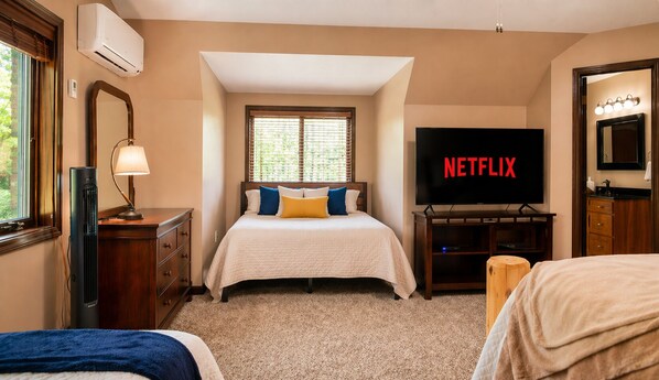 A hotel room with a bed, a TV displaying 'NETFLIX,' a dresser, a lamp, and a window with blinds. - Peaceful Lakefront Lodge near Pictured Rocks –with Kayaks, Firepit and Dock (Wetmore)