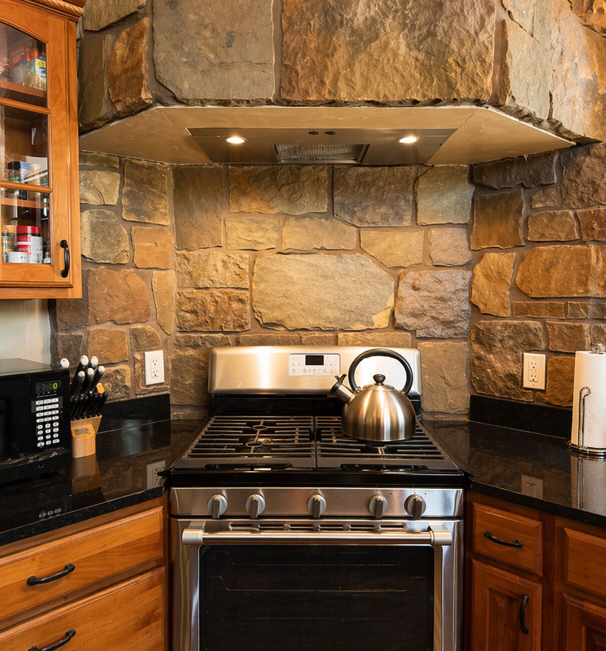 A kitchen with a stone wall, a stainless steel stove, and wooden cabinets.
