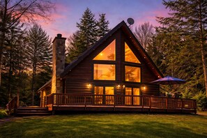 A log cabin with a stone chimney, large windows, and a deck with outdoor seating. - Peaceful Lakefront Lodge near Pictured Rocks –with Kayaks, Firepit and Dock (Wetmore)