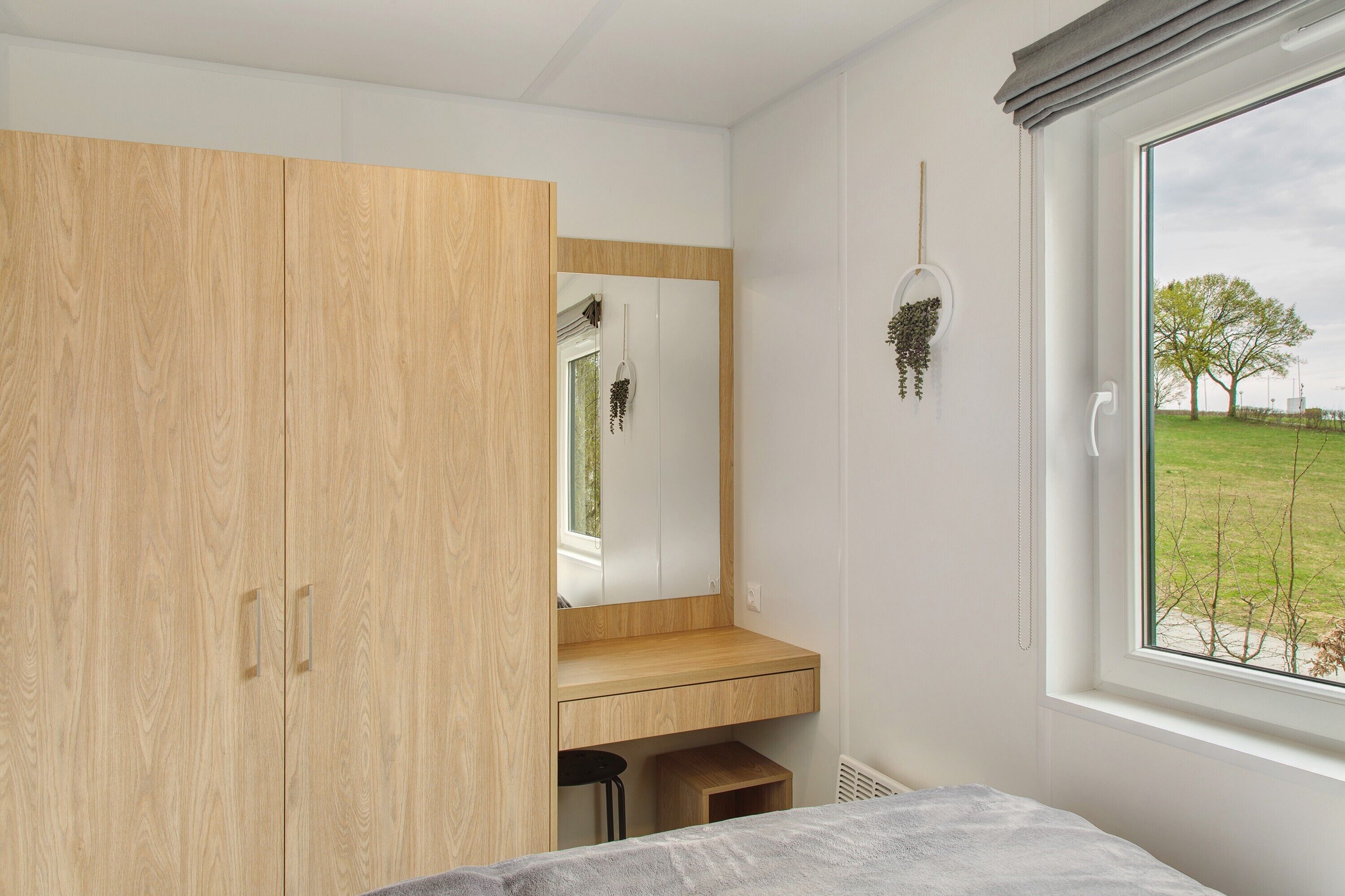 Un dormitorio con un armario de madera, un espejo, un taburete pequeño y una ventana con vistas a un campo cubierto de hierba.