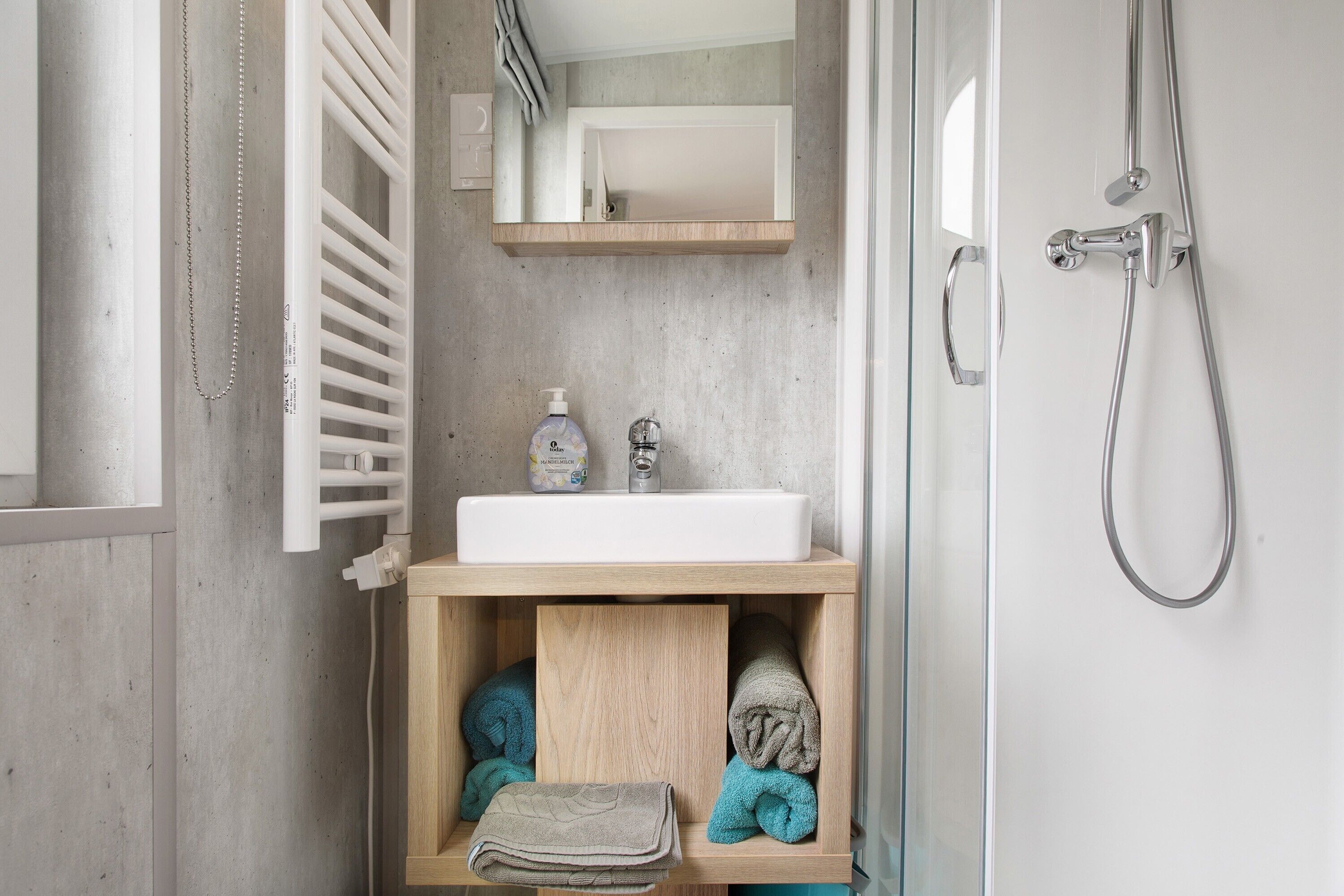 Ein Badezimmer mit weißem Waschbecken, Holz-Badmöbeln, Handtuchhalter und einer Dusche.
