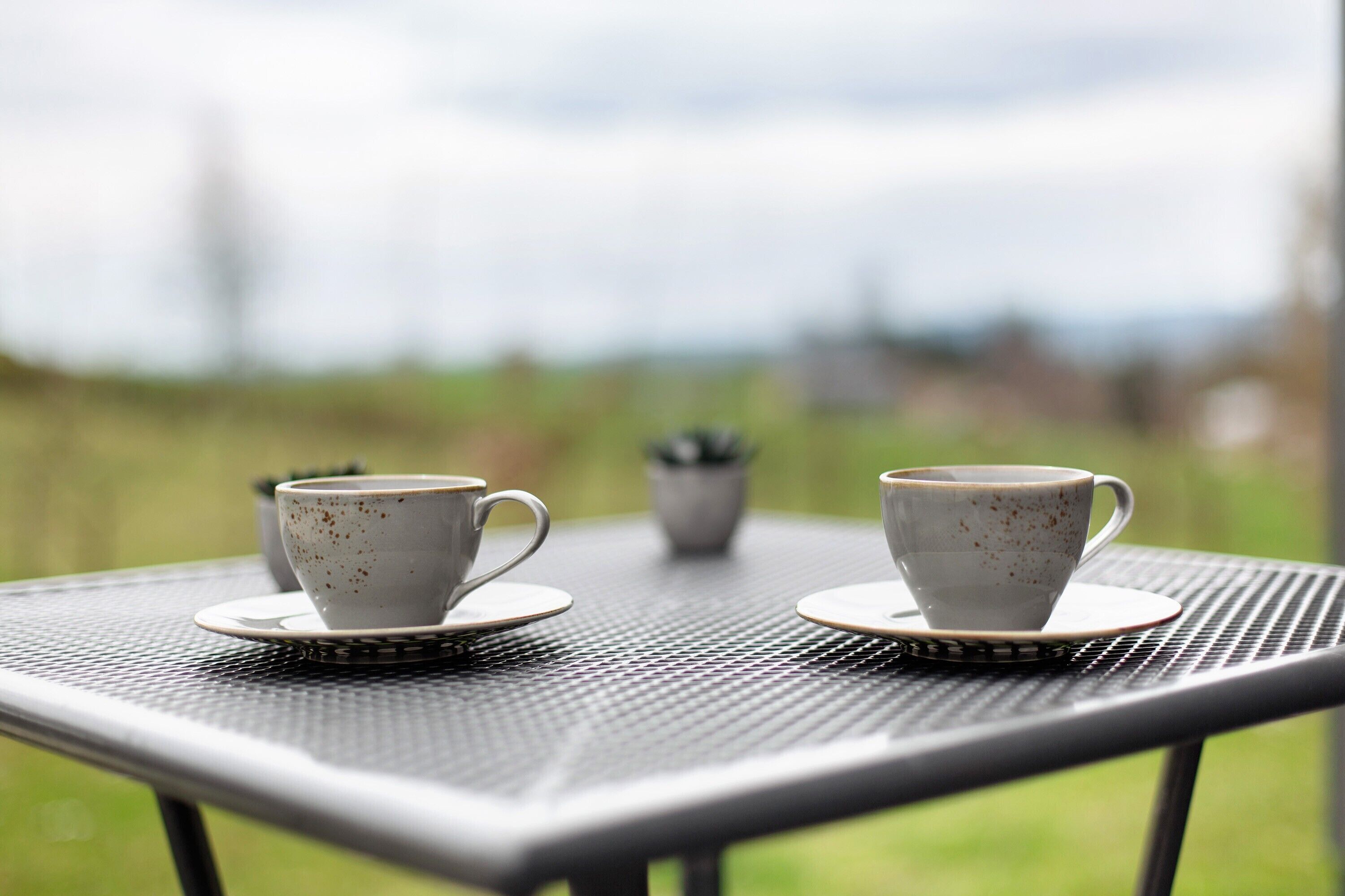 Two white cups on a metal table with a blurred outdoor background.