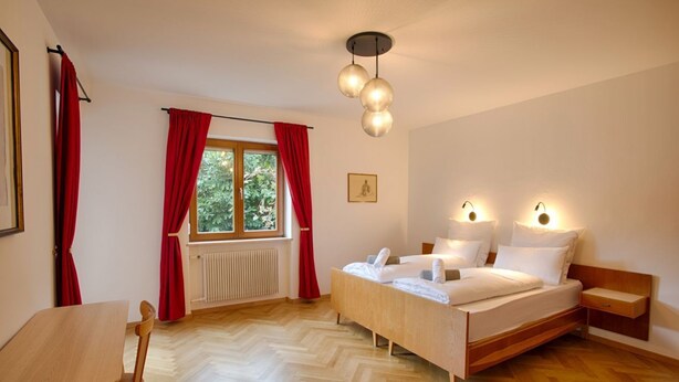 A bedroom with a wooden bed, a wooden table, and a chair. There is a window with red curtains, a radiator, and a picture on the wall. - Apartment Hofgarten - Alpstay (Naturno)