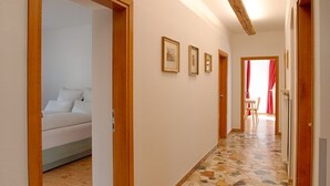 A hallway with a bed in the left room, a dining area with a table and chairs, and a door leading to another room. - Apartment Hofgarten - Alpstay (Naturno)