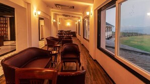 A corridor with leather chairs and tables, a large window with a view of a building and a grassy area, and a ceiling with recessed lighting. - Royal gold feildd club resorts (Dapoli)