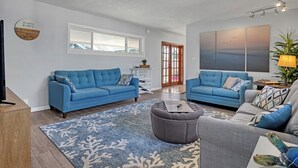 Ein geräumiges Wohnzimmer mit zwei blauen Sofas, einem grauen Sessel und einem grauen Hocker. Es gibt ein großes Fenster, ein Bild an der Wand und eine Pflanze in der Ecke.