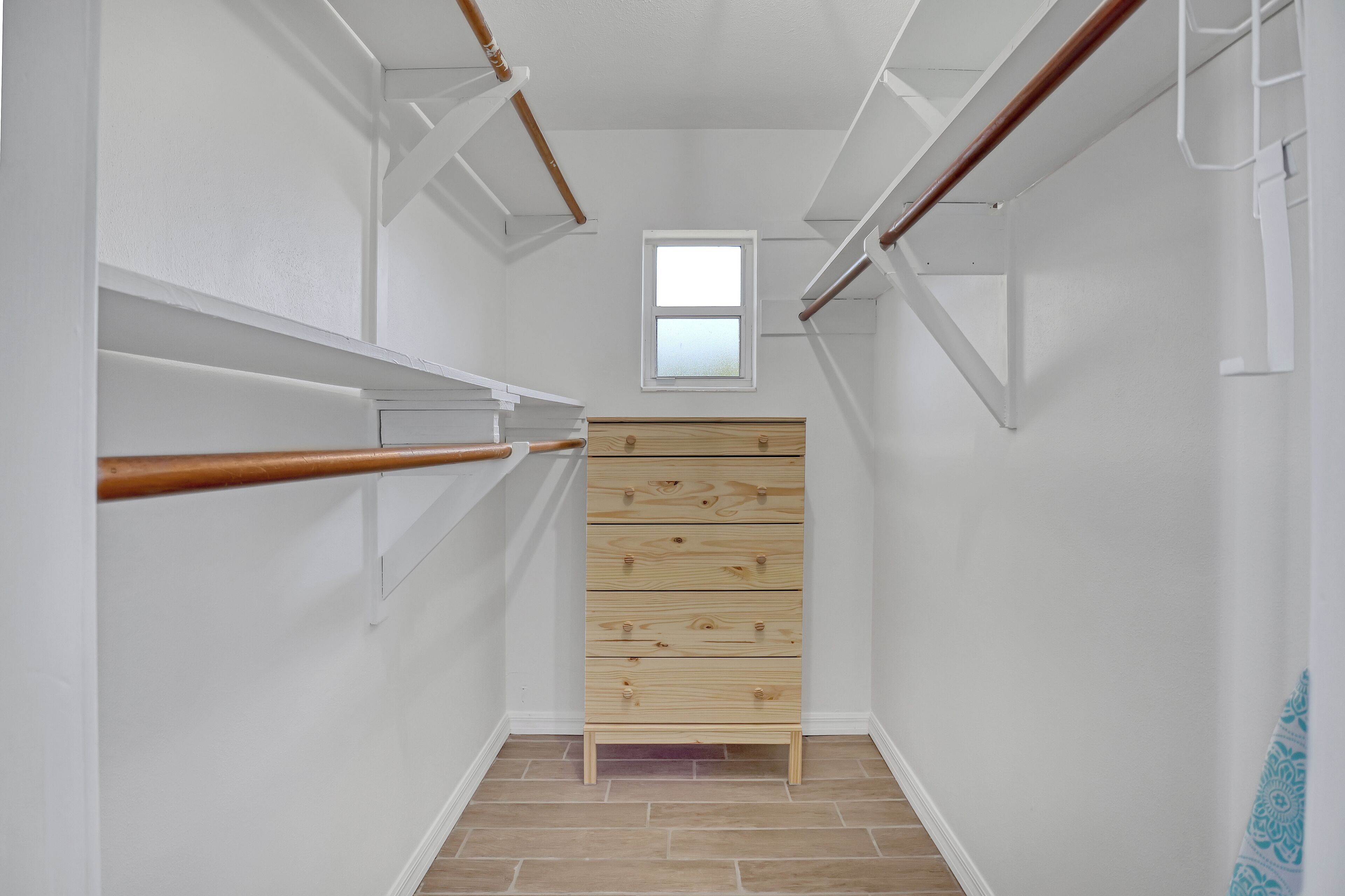 A narrow hallway with white walls, a wooden dresser, and a small window.