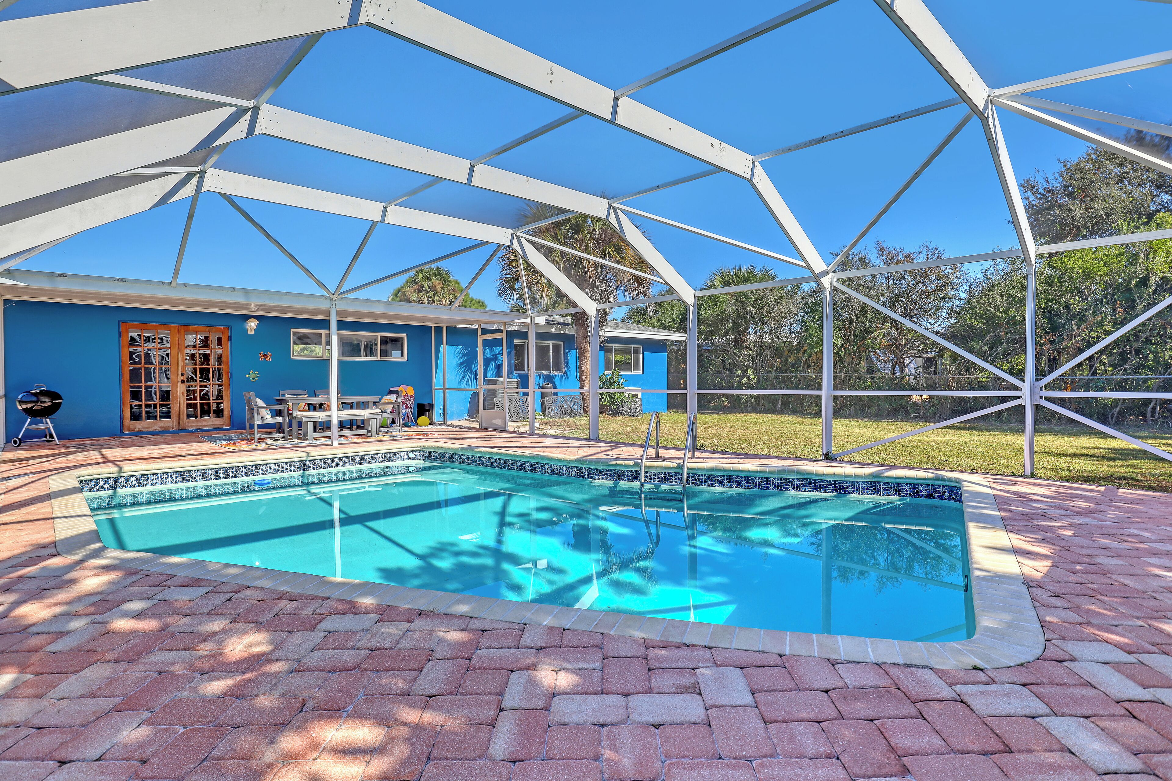 A swimming pool with a covered patio area, a blue building, and a grassy lawn in the background.