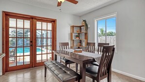 Ein Essbereich mit Holztisch, Stühlen und einer Bank. Eine Deckenventilator, ein Fenster mit Blick auf einen Pool sowie ein Holzregal mit Dekorationen sind ebenfalls vorhanden.