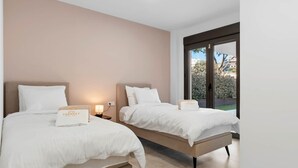 Una habitación doble con dos camas, cada una con cubrecama y almohadas blancas, una mesita de noche con lámpara y vistas al exterior a través de una puerta de vidrio.