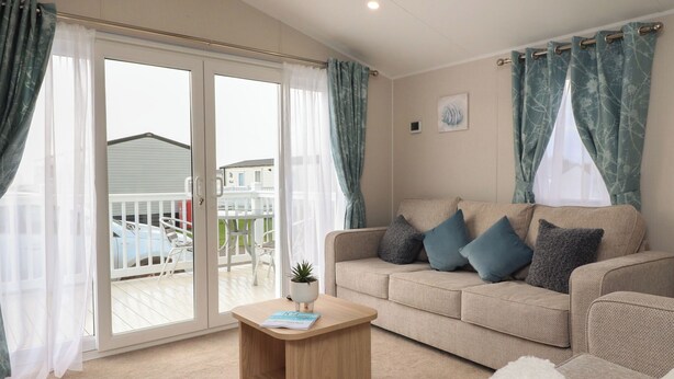 A living room with a beige sofa, a wooden coffee table, and a view of a balcony with outdoor furniture. - Poppy Lodge (Morecambe)