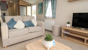 A living room with a beige sofa, a coffee table with a plant, a TV on a wooden stand, and a window with curtains. - Poppy Lodge (Morecambe)