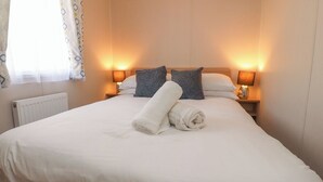 A neatly made bed with white linens, two pillows, and a rolled towel. There are two bedside lamps with lampshades, a window with a curtain, and a radiator under the bed. - Poppy Lodge (Morecambe)