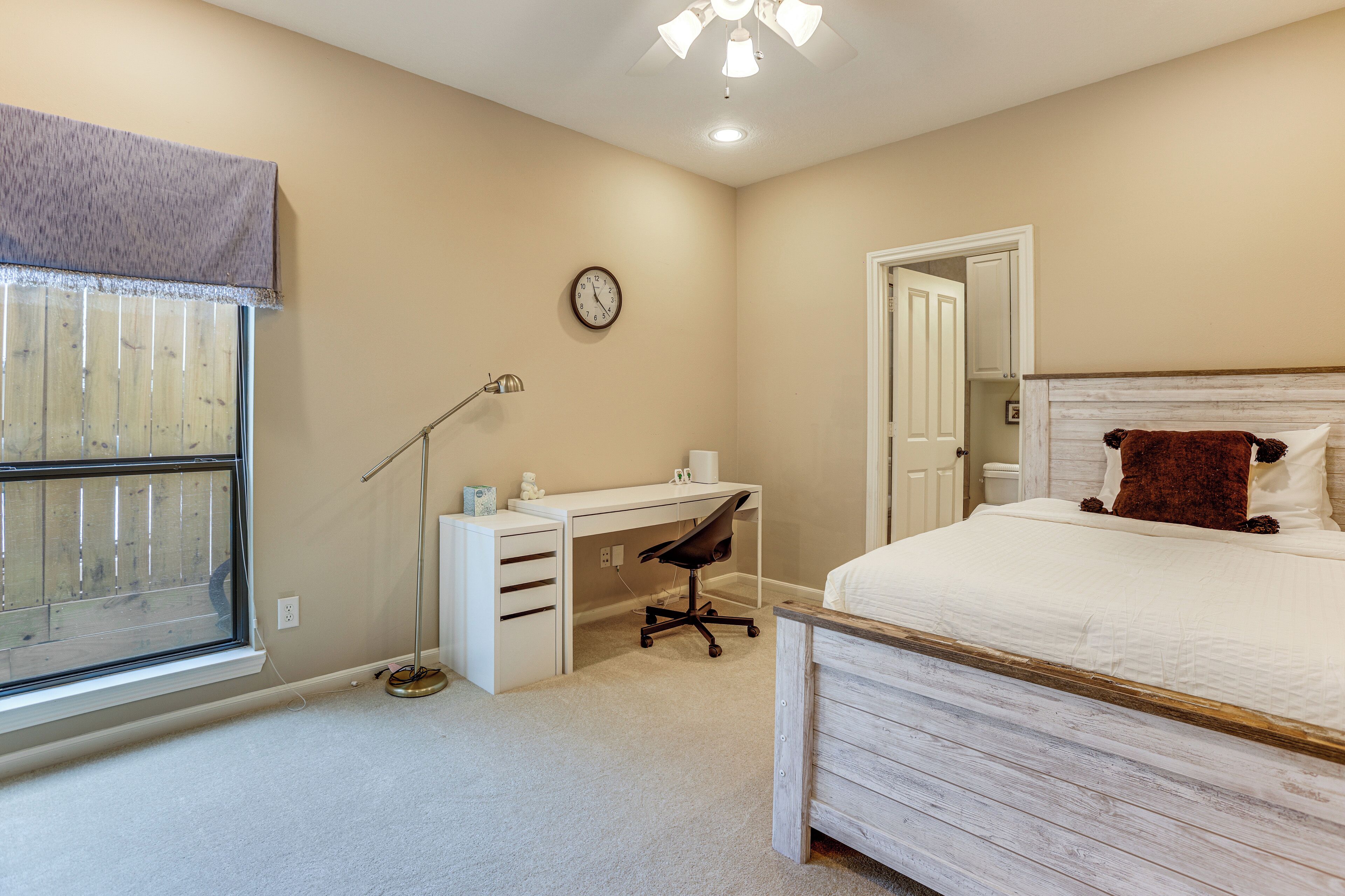 A bedroom with a bed, a desk, a chair, a window with blinds, a clock, and a door.