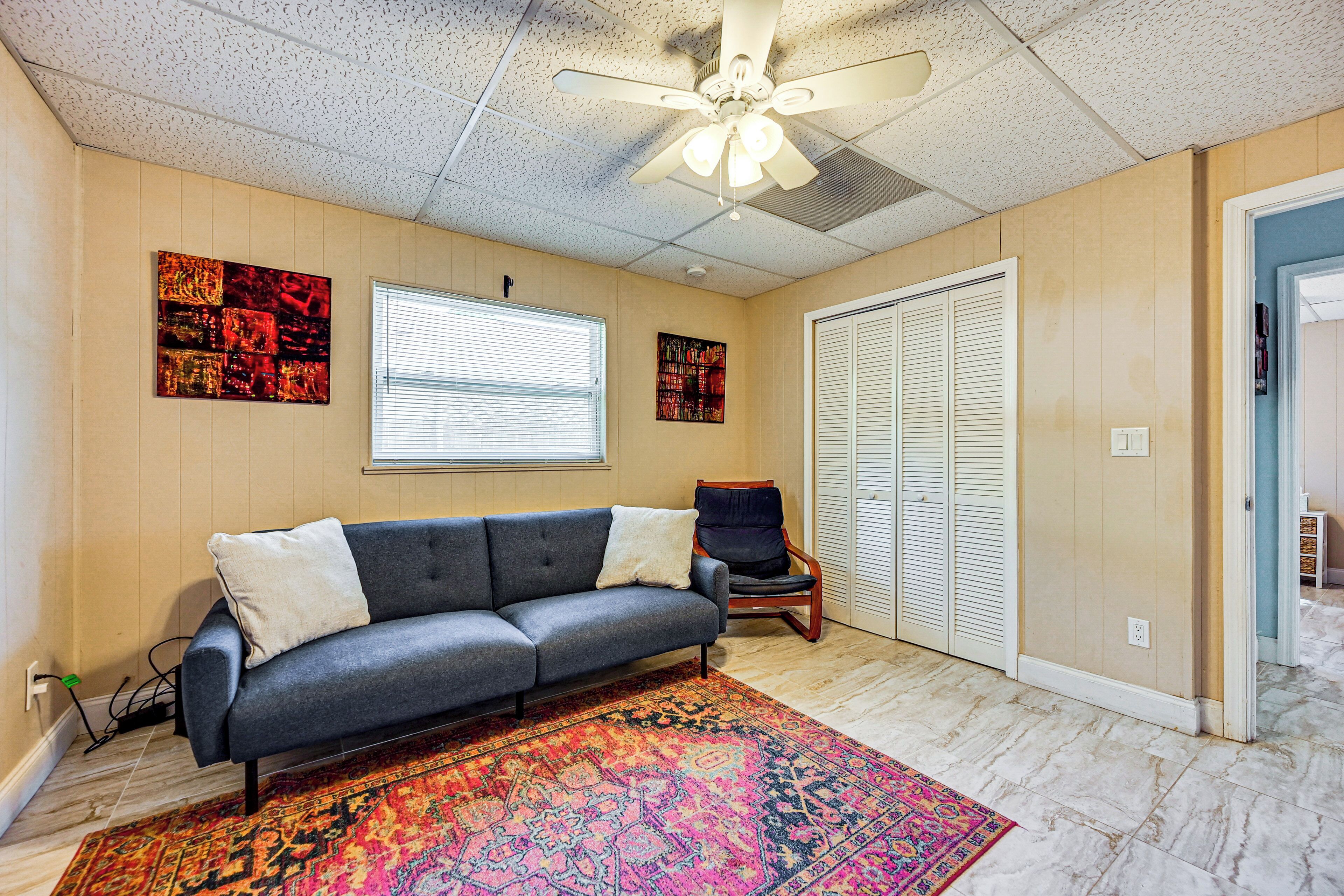 A room with a gray sofa, a colorful rug, a ceiling fan, and a window with blinds.
