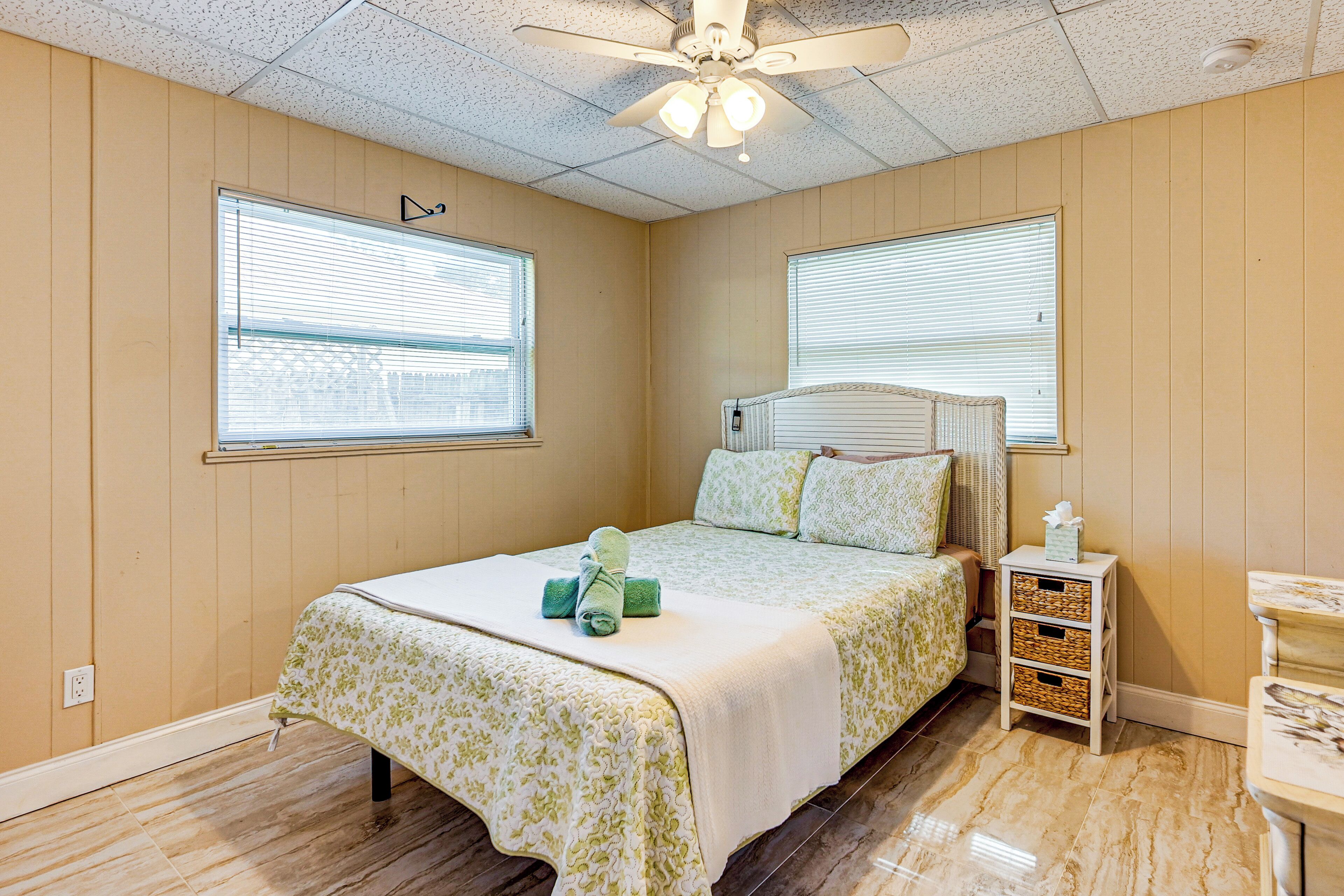 A bedroom with a bed, two windows, a ceiling fan, and a bedside table with a basket.