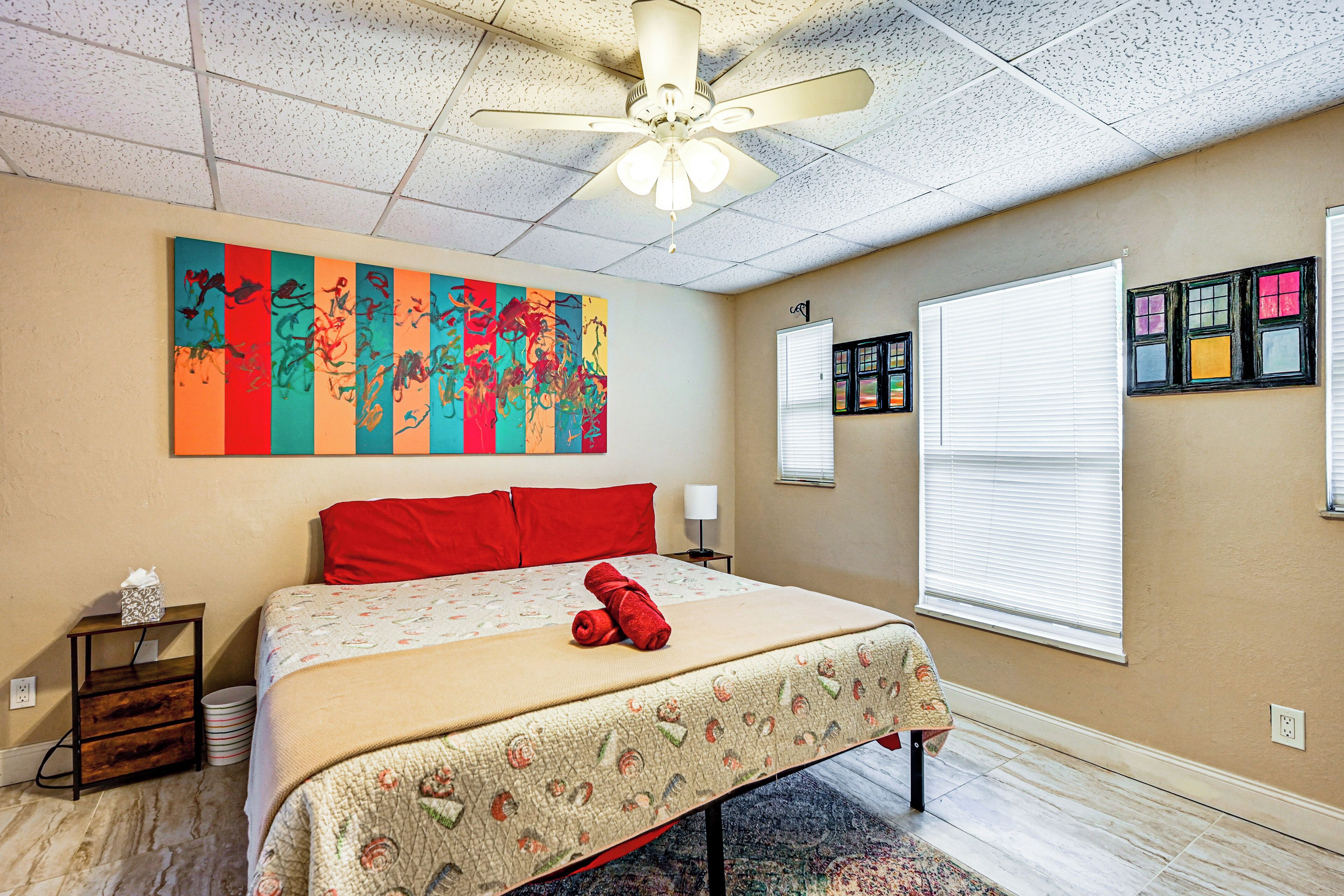A hotel room with a bed, a ceiling fan, a colorful abstract painting, a window with blinds, and a small table with a lamp.