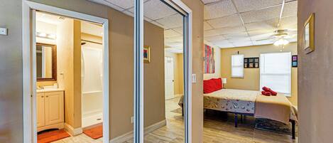 A room with a sliding door leading to a bathroom, a bed with a red pillow, and a ceiling fan.