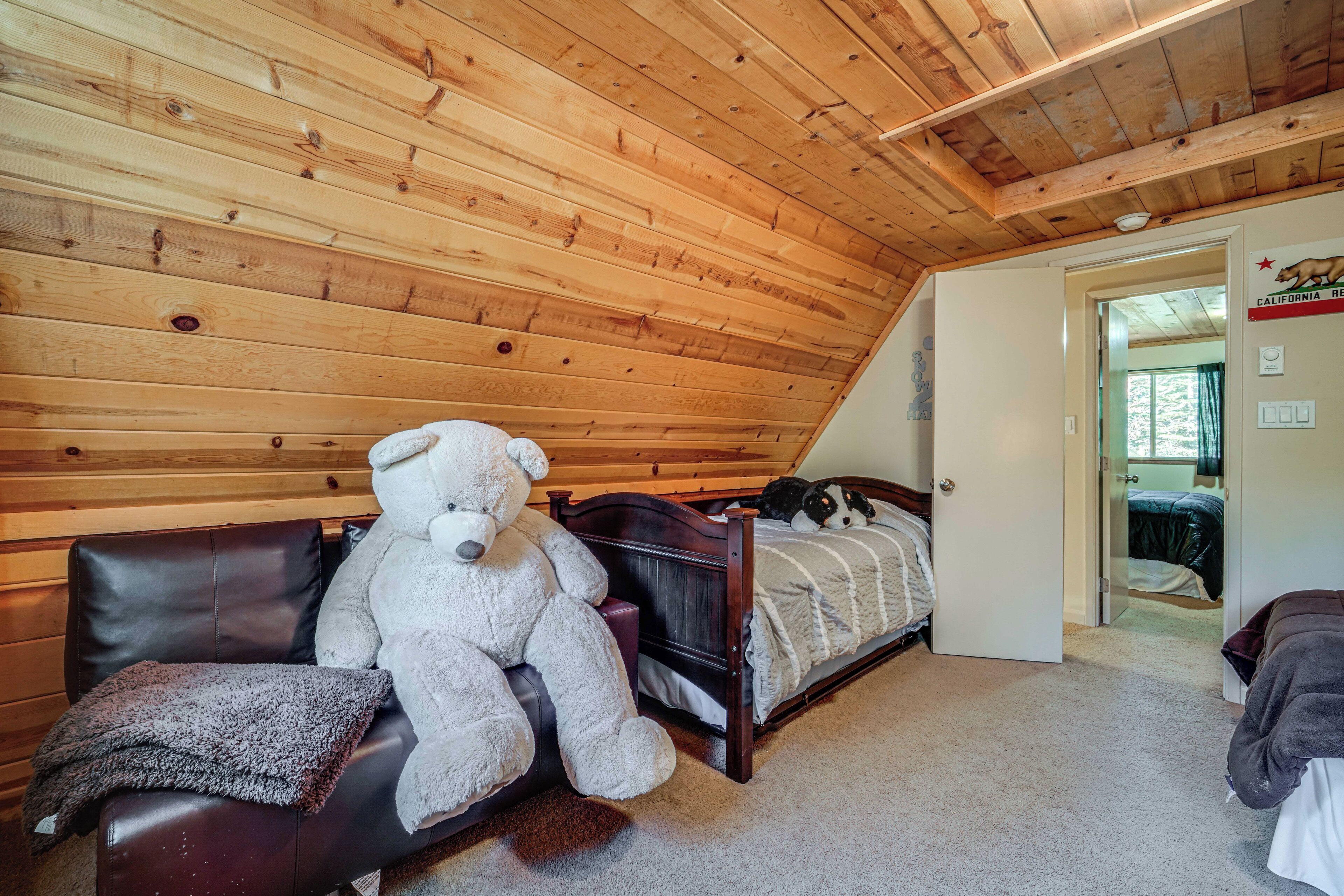 Una habitación con techo de madera, una cama con cubrecama blanco, un gran oso de peluche en una silla de cuero y una puerta que da a otra habitación.