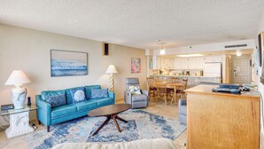 A living room with a blue sofa, a coffee table, and a rug. There is a kitchen in the background with white cabinets and a dining area. - Steps to Shore, Pool Access: New Smyrna Beach Gem! (New Smyrna Beach)