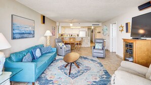 A living room with a blue sofa, a coffee table, and a TV on a wooden cabinet. There is a kitchen in the background with a dining area. - Steps to Shore, Pool Access: New Smyrna Beach Gem! (New Smyrna Beach)