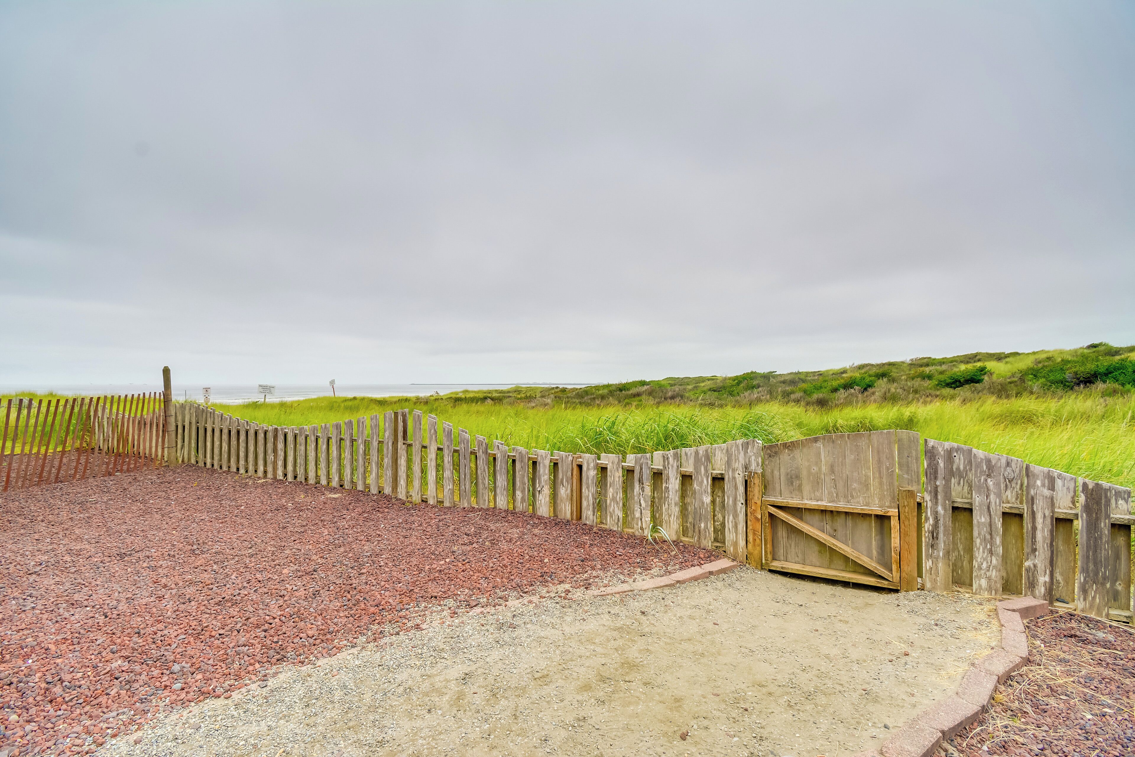 Un área de playa de grava con una valla de madera y una puerta, rodeada de dunas cubiertas de hierba.
