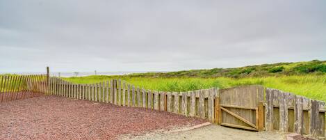 Un área de playa de grava con una valla de madera y una puerta, rodeada de dunas cubiertas de hierba.