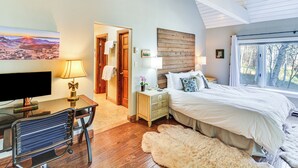Ein Schlafzimmer mit einem Bett, einem Schreibtisch mit Computer, einem Stuhl, einer Holzverkleidung an der Wand und einem Fenster mit Blick auf Bäume.