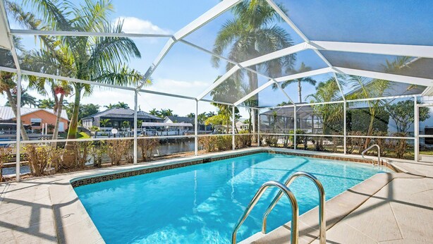 A clear blue pool with a metal ladder, surrounded by a covered area with palm trees and buildings in the background. - New Listing! Canalfront Comfort | Heated Pool, Patio & Palm-Lined Views (Cape Coral)