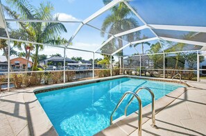 A clear blue pool with a metal ladder, surrounded by a covered area with palm trees and buildings in the background.