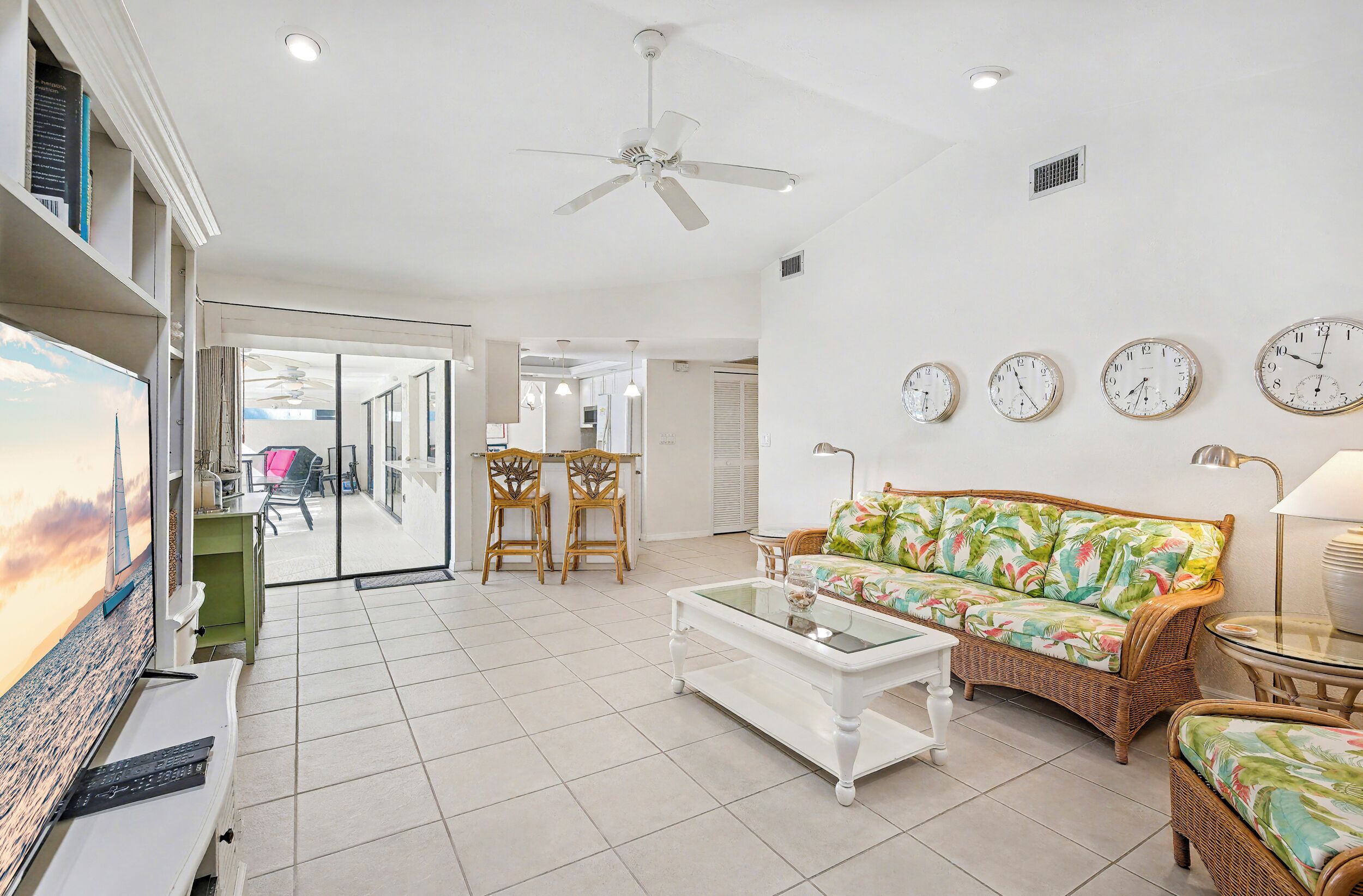 A spacious living area with a sofa, coffee table, and a television. There are multiple clocks on the wall and a ceiling fan.