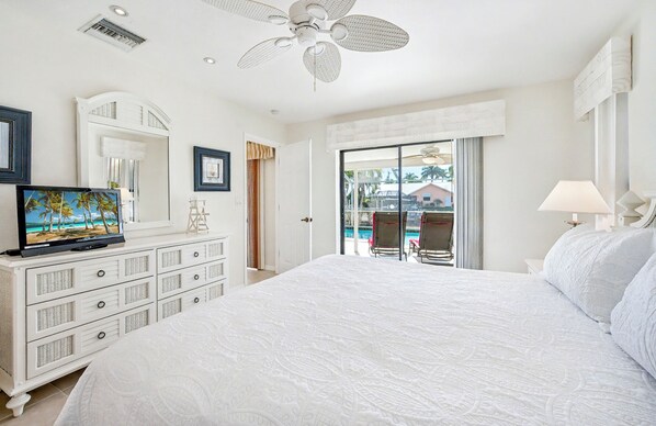 A spacious bedroom with a large bed, a ceiling fan, a television, and a view of a pool area.