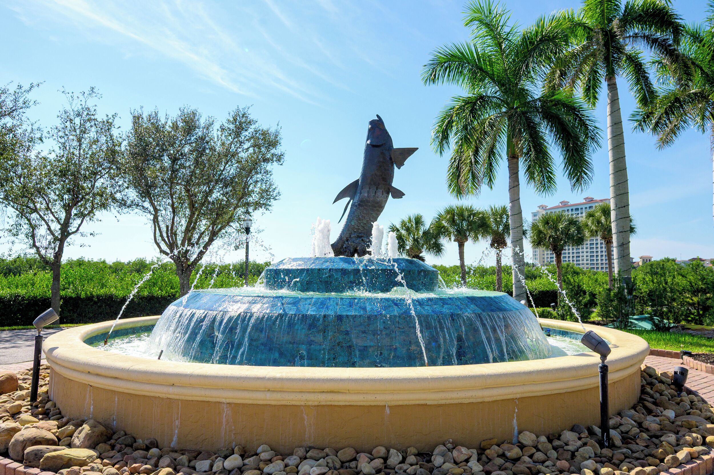 A fountain with a fish sculpture in the center, surrounded by palm trees and a clear sky.