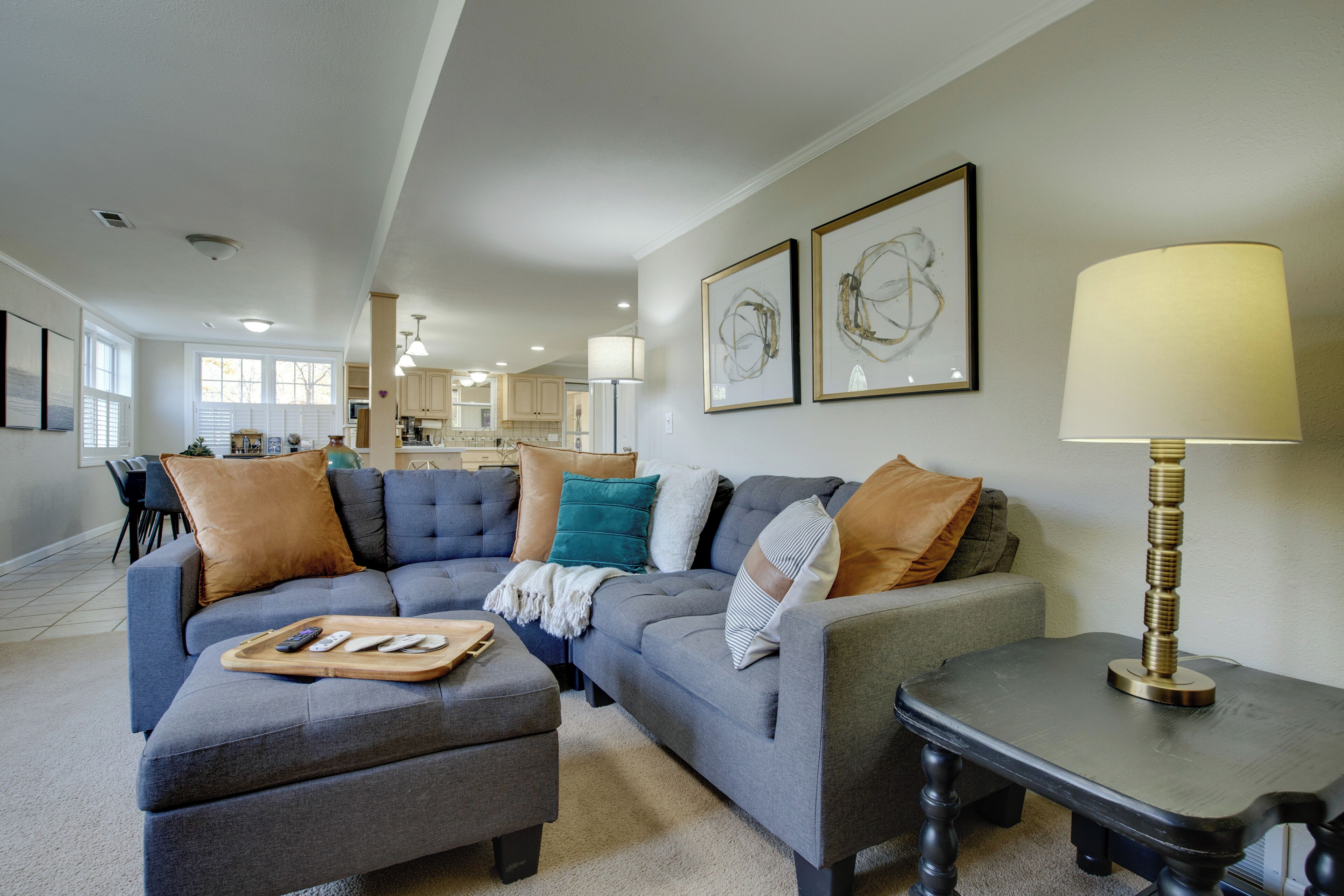 A modern living room with a sectional sofa, ottoman, and a side table with a lamp. There are framed artworks on the wall and a kitchen area in the background.