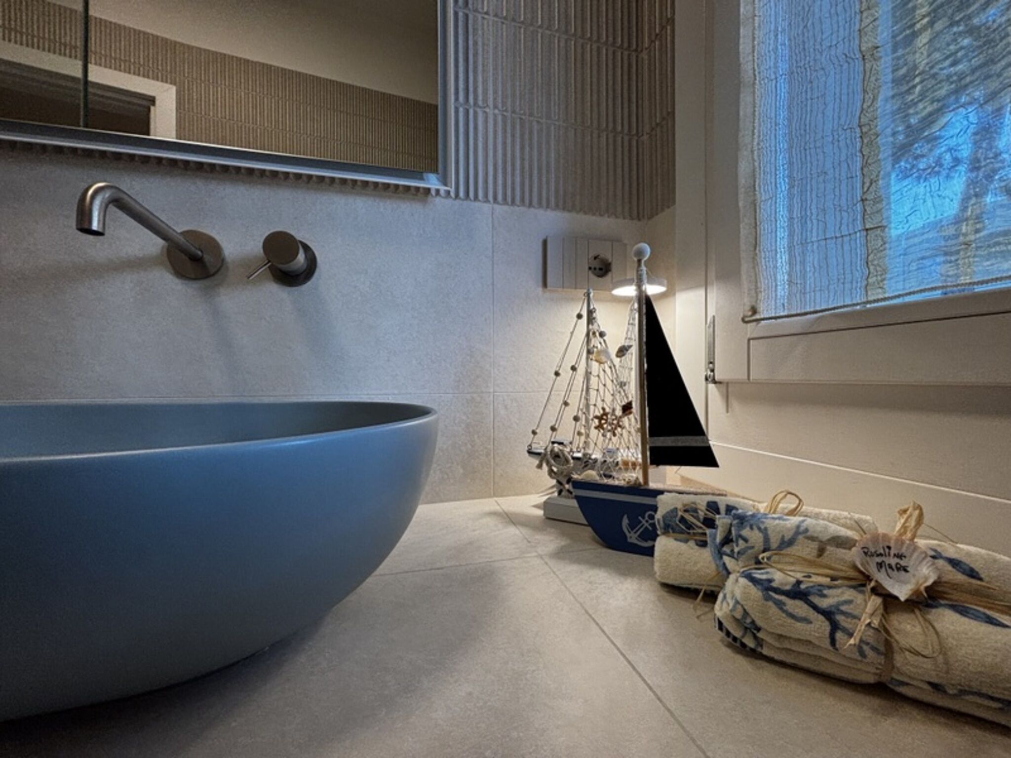 Un baño moderno con un lavamanos azul grande, un modelo de barco y una ventana con vistas a árboles.