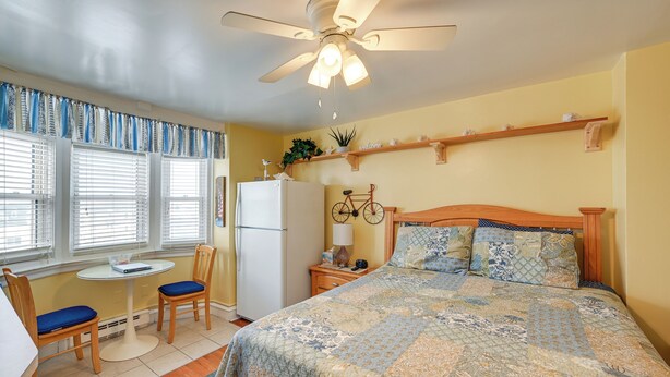 Ein Schlafzimmer mit Bett, Deckenventilator, Fenster mit Jalousien, kleinem Tisch mit Stühlen, Nachttisch und einer Kommode mit Dekoration.