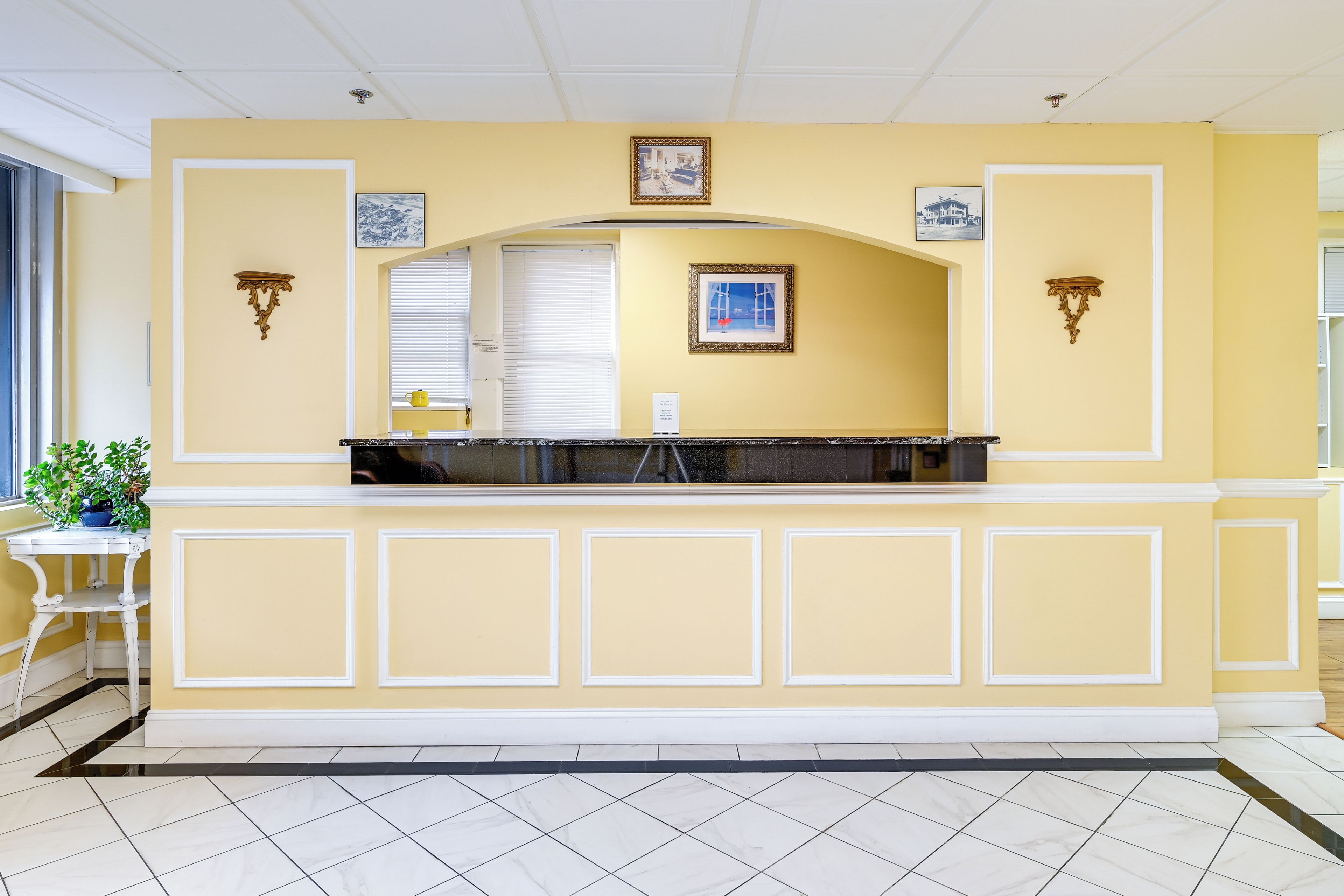 A reception area with a yellow wall, white trim, and a tiled floor. There are two wall-mounted light fixtures and a framed picture above the reception desk.