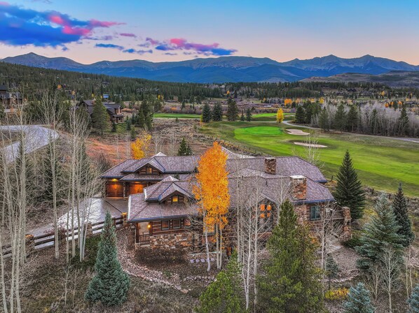 A large, rustic lodge with a stone exterior and multiple chimneys, surrounded by a forested area and a golf course. - Luxury Home, Massive Views, Pool Table, Hot Tub, Nordic Skiing and Golf (Breckenridge)