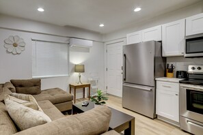 Una cocina moderna con armarios blancos, un refrigerador de acero inoxidable y un horno. Hay un comedor con un sofá marrón y una pequeña mesa.