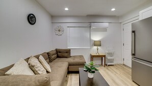 A modern living room with a sectional sofa, a small table with a plant, a wall clock, and a refrigerator. - 10 Mi to Riverside! Apt Near Ruhs Medical Center (Moreno Valley)