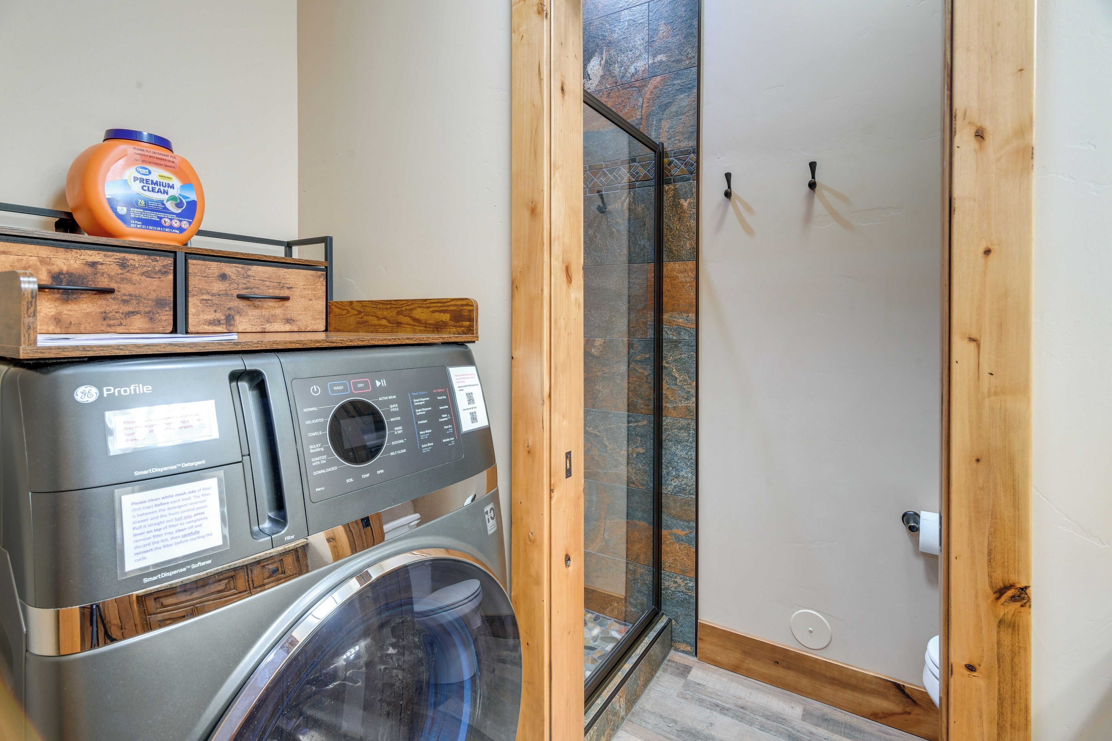 Un espace buanderie compact comprenant un lave-linge et un sèche-linge à chargement frontal, un meuble de rangement en bois et une cabine de douche en verre.