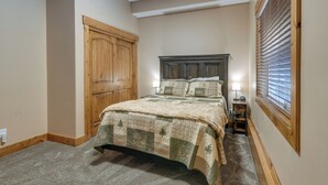 A bedroom with a large bed, a wooden wardrobe, a small bedside table with a lamp, and a window with blinds. - Gateway to Yellowstone! Modern Island Park Cabin (Island Park)