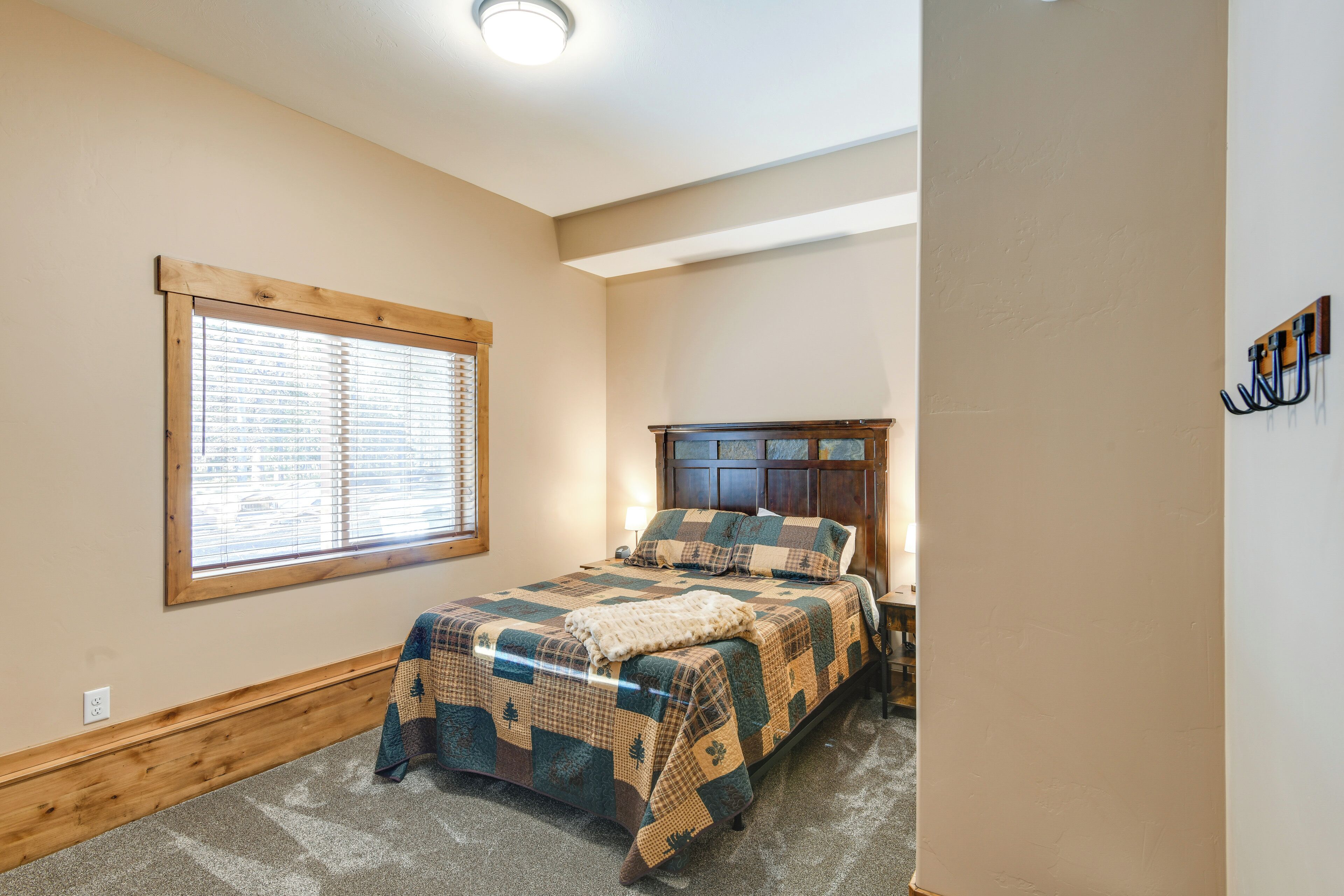 A bedroom with a wooden headboard, a quilted bedspread, a window with blinds, and a wall-mounted coat rack.