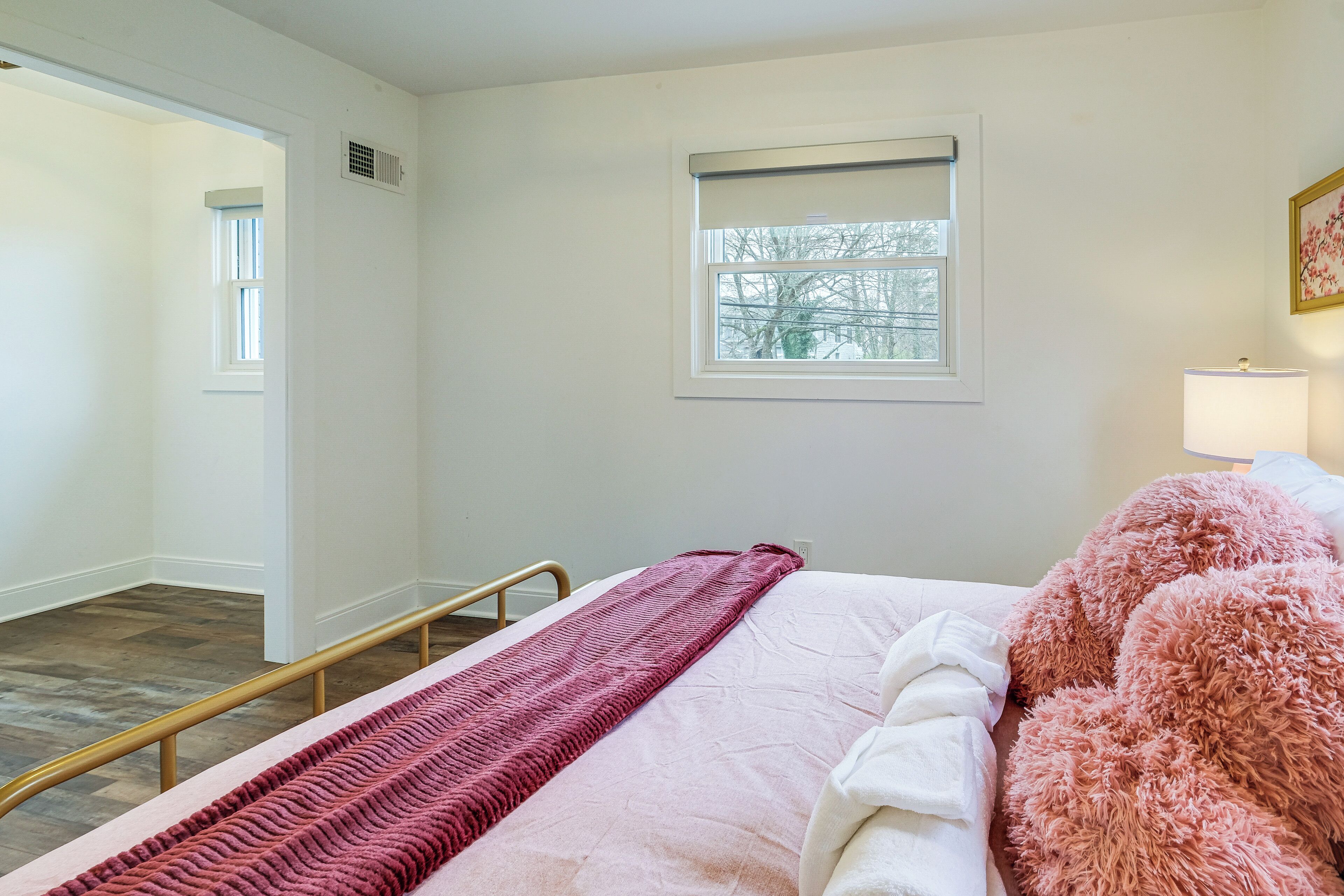 A bedroom with a bed, a window, a bedside lamp, and a door leading to another room.